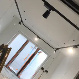 Czarna szyna oświetleniowa z reflektorami punktowymi zamontowana na suficie w trakcie prac wykończeniowych, okno zabezpieczone folią, drabina i lampa budowlana widoczne w pomieszczeniu.