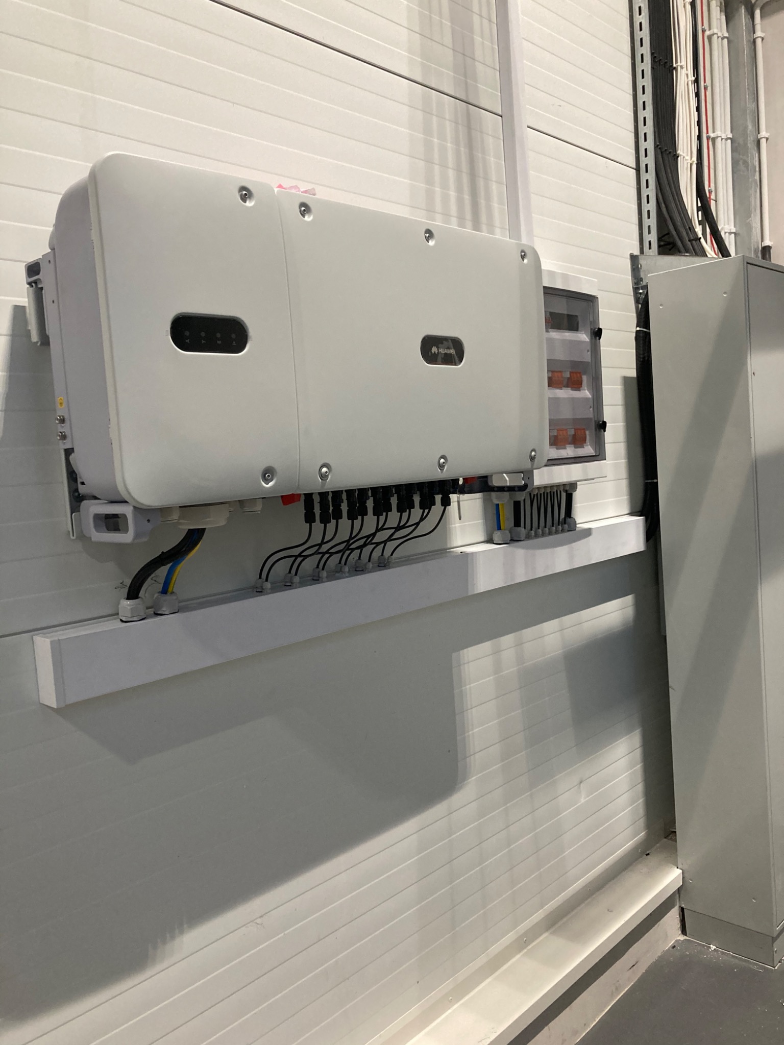 Biały falownik marki Huawei zamontowany na ścianie z blachy trapezowej, widoczne kable i skrzynka elektryczna w tle.