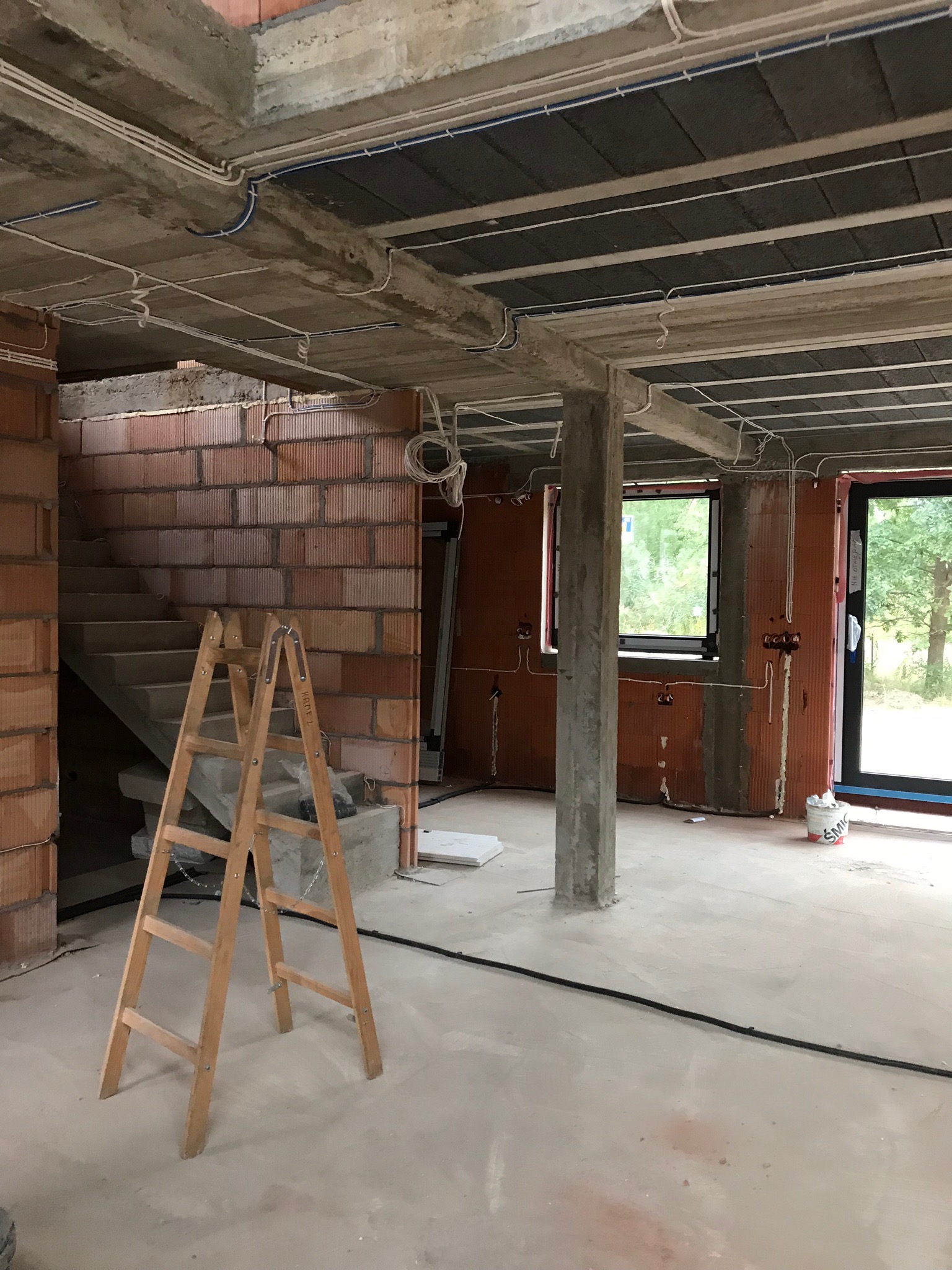 Wnętrze w trakcie budowy z widoczną instalacją elektryczną pod sufitem, ceglanymi ścianami i drewnianą drabiną opartą o ścianę ze schodami.