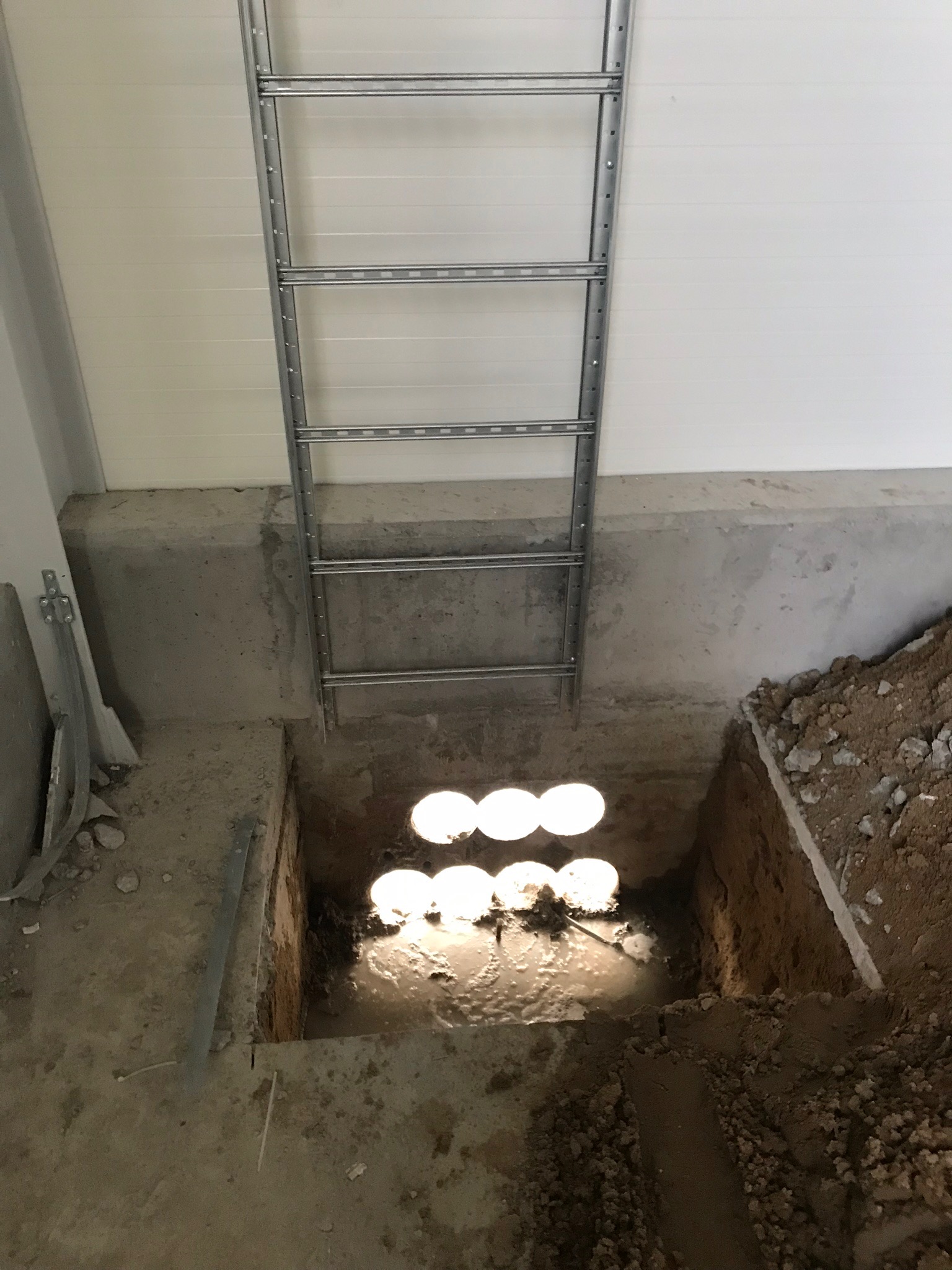 Wykop w betonowej posadzce z widocznymi przewodami w wodzie, drabina oparta o ścianę, surowy stan wykończenia.