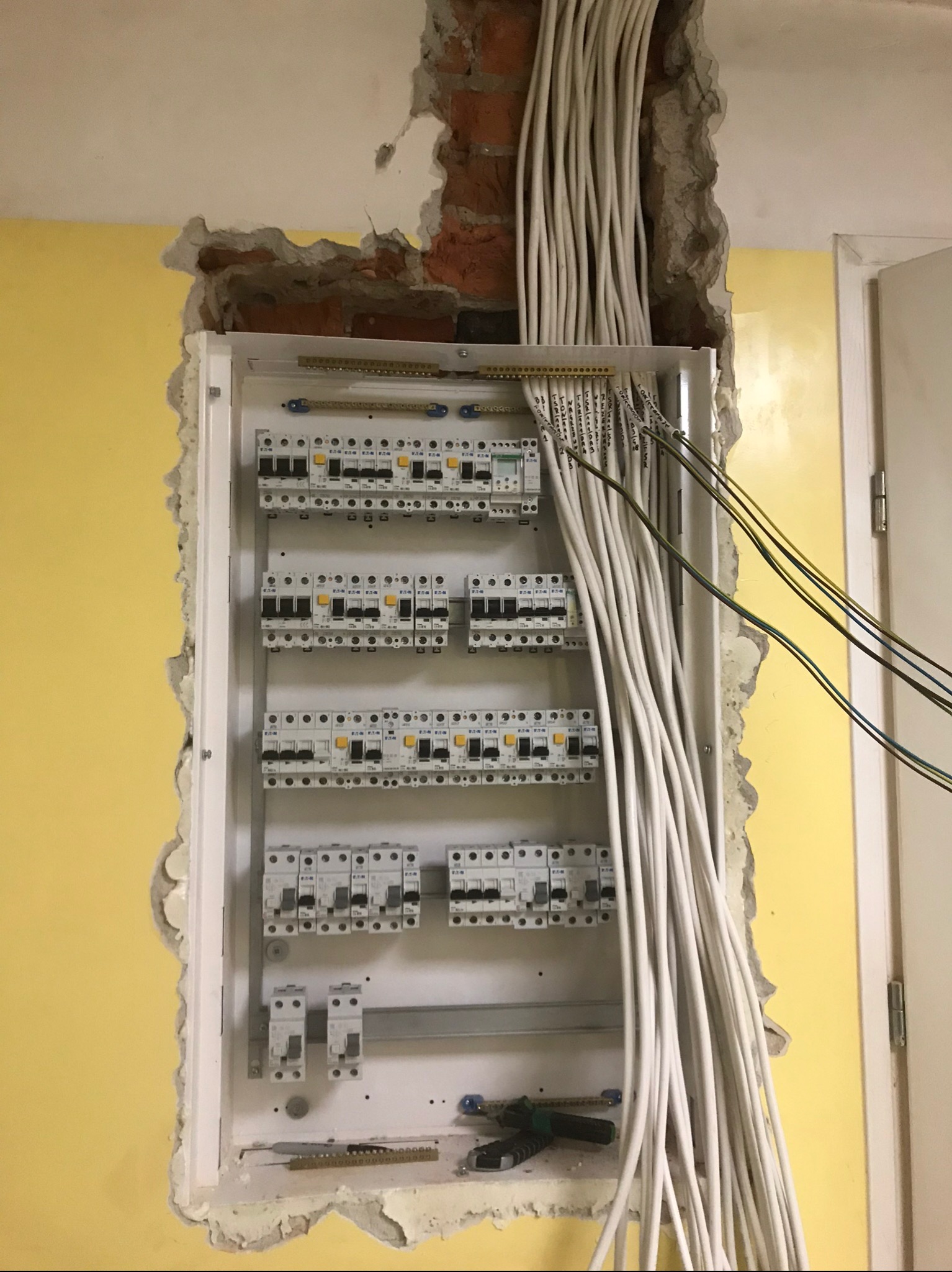 Rozdzielnica elektryczna z licznymi przewodami wystającymi ze ściany, widoczne uszkodzenia tynku i cegły wokół obudowy, żółta ściana w tle.