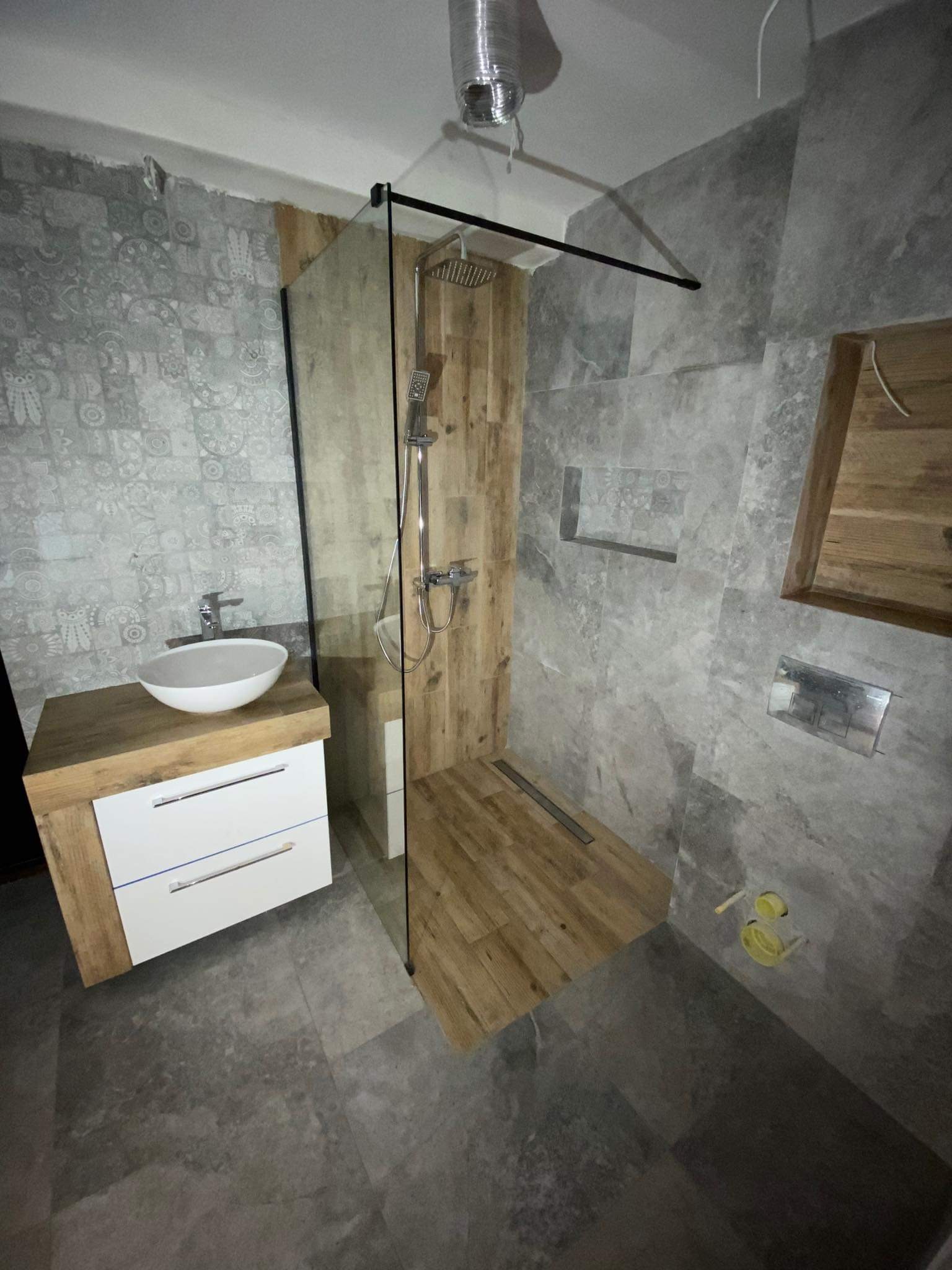 Nowoczesna łazienka z kabiną prysznicową typu walk-in wykończoną drewnem, umywalką nablatową na szafce z białymi szufladami oraz szarą płytką imitującą kamień na ścianach i podłodze.