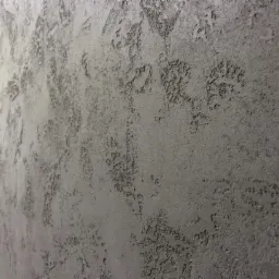 Szara ściana z dekoracyjną strukturą, widoczne fragmenty schodów w lewym dolnym rogu, nierównomierne oświetlenie.