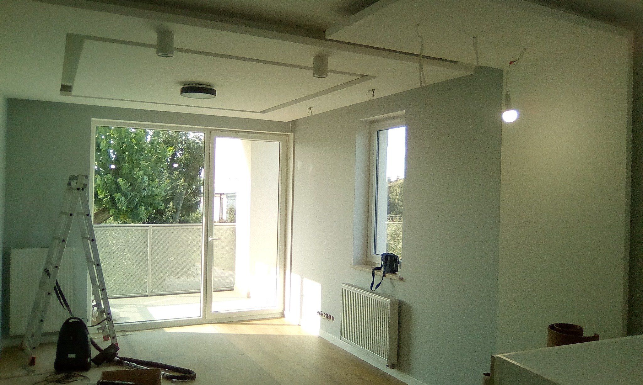 Wnętrze jasnego pokoju w trakcie prac wykończeniowych, widoczna drabina oparta o ścianę, odsłonięte kable, sufit z dekoracyjnymi elementami, parkiet na podłodze i grzejnik pod oknem.
