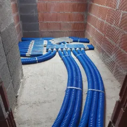 Niebieskie rury wentylacyjne z metalowymi obejmami, rozdzielacz powietrza w surowym wnętrzu z ceglanymi ścianami podczas montażu.