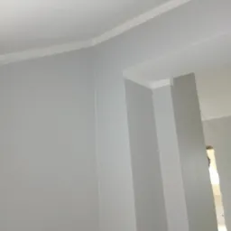 Świeżo pomalowane na szaro ściany i sufit w pomieszczeniu z białymi listwami dekoracyjnymi przy suficie i ościeżnicą drzwiową.