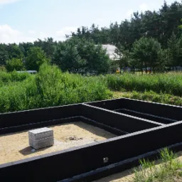 Zalane, czarne fundamenty domu jednorodzinnego na piaszczystym podłożu, widoczne bloczki betonowe przygotowane do dalszych prac, tło z zielenią i drzewami.