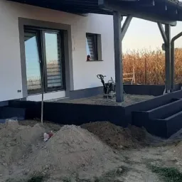 Przygotowanie podłoża pod taras z kostki przy domu jednorodzinnym, widoczna ubijarka wibracyjna i pryzma piasku.