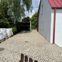 Ułożona kostka granitowa prowadząca do nowoczesnego domu z czerwoną dachówką, obok ułożone worki z materiałem na paletach.