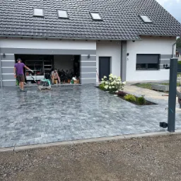 Układanie szarej kostki na podjeździe domu jednorodzinnego, pracownik używa zagęszczarki, drugi odpoczywa w garażu, widoczne białe hortensje i taczka z piaskiem.