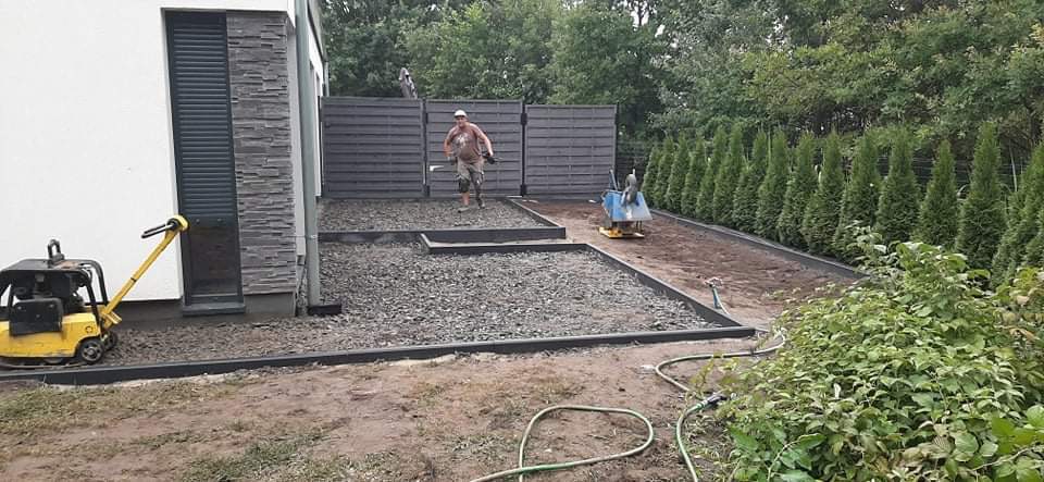 Przygotowanie podłoża pod ścieżkę z kostki: wyrównany teren, obrzeża, zagęszczarka w tle, pracownik rozrzucający kamień