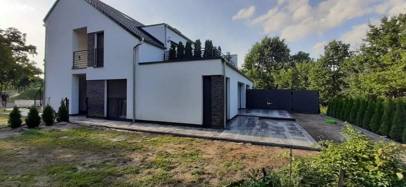 Nowoczesny dom jednorodzinny z szarą kostką brukową na tarasie i ścieżce, widoczne nasadzenia iglaków i trawa, szare ogrodzenie w tle.