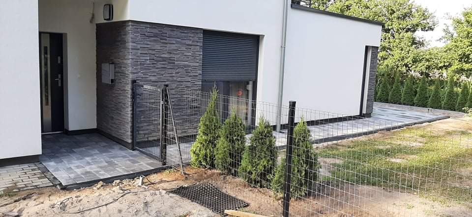 Ułożona szara kostka brukowa przy nowoczesnym domu z szarą elewacją, widoczny fragment metalowego ogrodzenia i posadzone tuje.