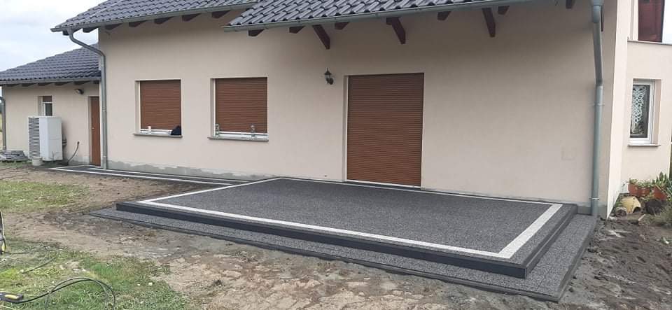 Wykończony taras z szarej kostki żywicznej z białym obramowaniem, prowadzący do drzwi domu z brązowymi roletami.