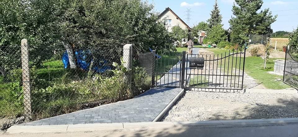 Ułożona ścieżka z szarej kostki prowadząca od furtki do domu z numerem 17, widoczny fragment ogrodu i siatka ogrodzeniowa.