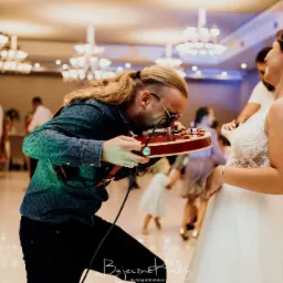 Muzyk z długimi włosami gra na gitarze elektrycznej, pochylając się nad nią podczas wesela, obok uśmiechnięta panna młoda w koronkowej sukni.