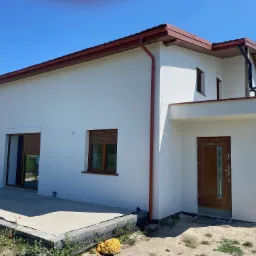 Nowo otynkowany, biały dom jednorodzinny z brązowymi elementami dachu i okien, widoczny z zewnątrz w słoneczny dzień.