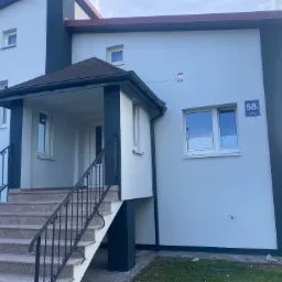 Dwukondygnacyjny dom z jasną elewacją, ciemnymi akcentami w postaci obramowań i kolumn, oraz schodami wejściowymi z ozdobną balustradą, widoczny numer domu 58a.