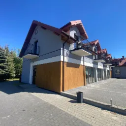 Szary budynek z balkonami i brązowym dachem w słoneczny dzień. Widok z dołu, ukazujący elewację i fragment parkingu z kostki brukowej. Częściowo widoczne drzewa.