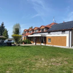 Dwukondygnacyjny budynek z panelami słonecznymi na dachu i nową elewacją w odcieniach brązu i szarości, otoczony zielonym trawnikiem w słoneczny dzień.