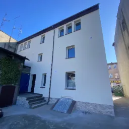 Odnowiona elewacja budynku z jasną fasadą, ciemnym dachem i nowymi oknami, widoczna z perspektywy podwórza w słoneczny dzień.