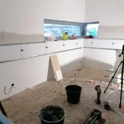 Przygotowanie do układania płytek: ściany z zamontowanymi profilami, narzędzia glazurnicze, poziomica na statywie, widok przez okno na zewnątrz.