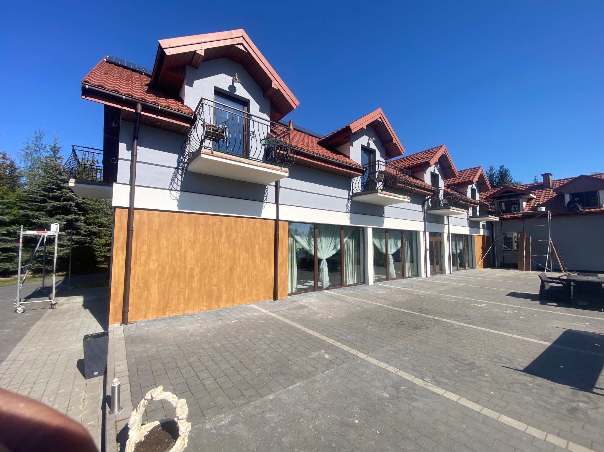 Odnowiony budynek z balkonami i czerwoną dachówką, widok z zewnątrz, jasne tynki i drewniane akcenty na elewacji. Błękitne niebo w tle.