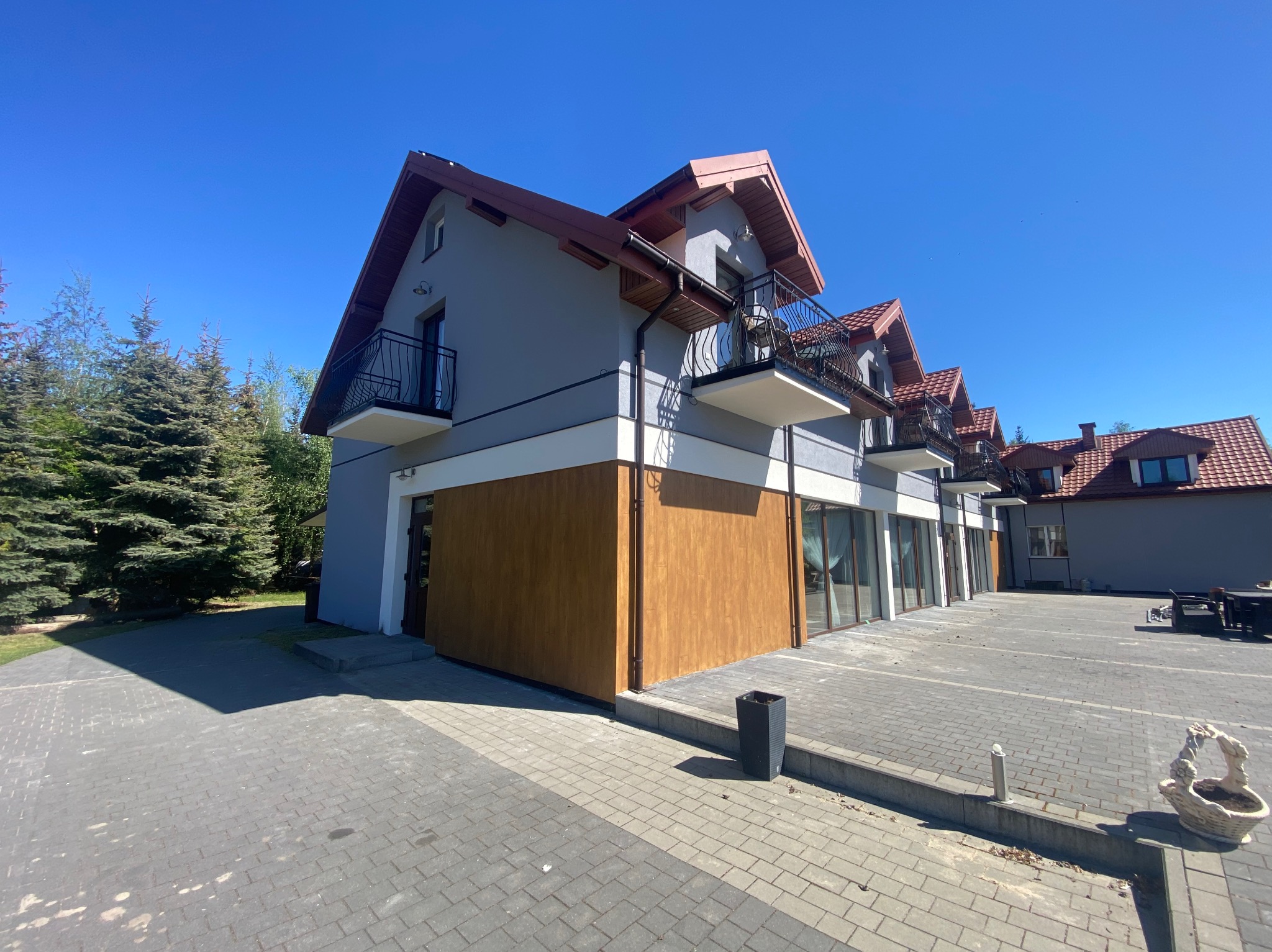 Szary budynek z balkonami i brązowym dachem w słoneczny dzień. Widok z dołu, ukazujący elewację i fragment parkingu z kostki brukowej. Częściowo widoczne drzewa.