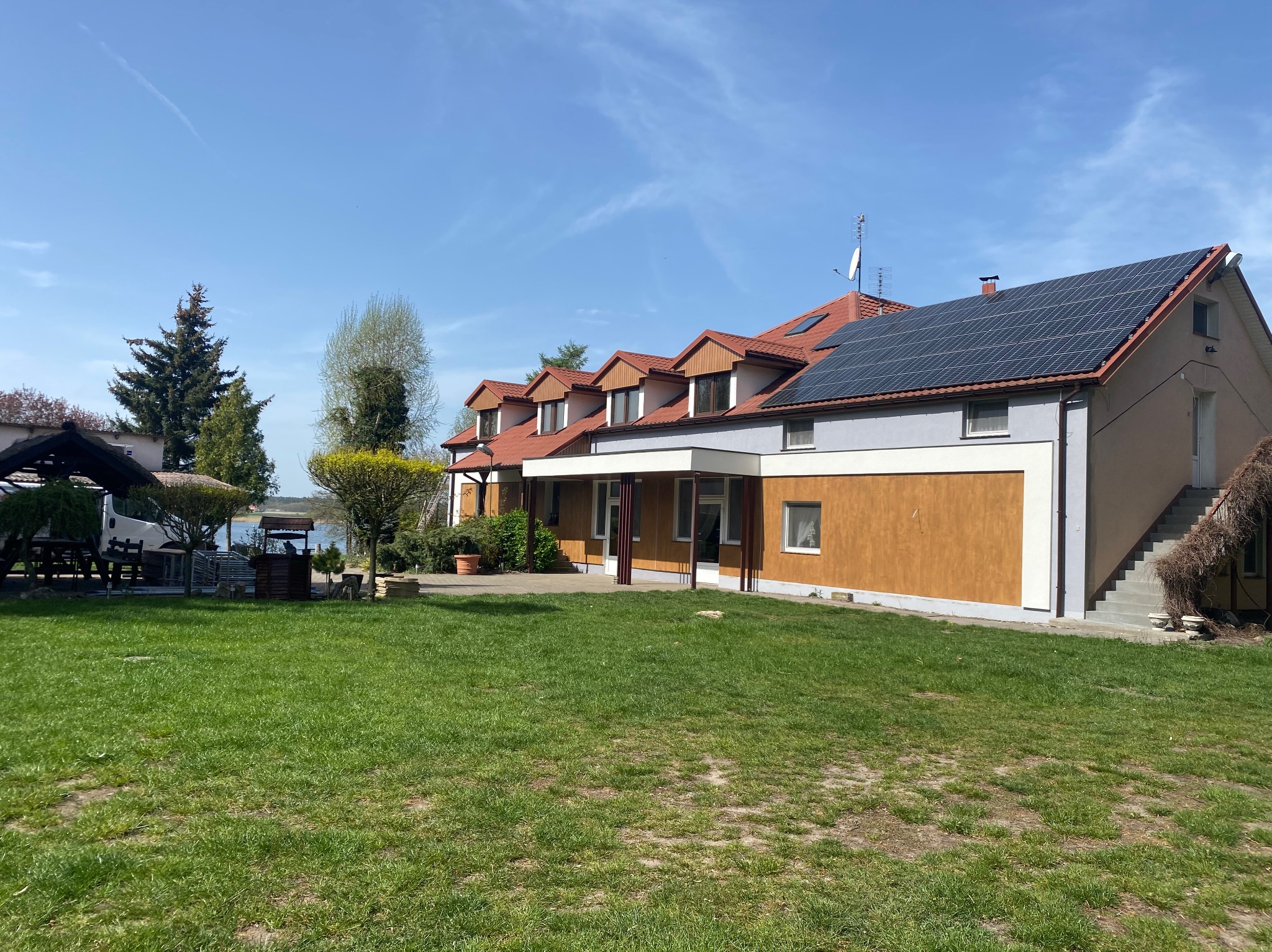 Dwukondygnacyjny budynek z panelami słonecznymi na dachu i nową elewacją w odcieniach brązu i szarości, otoczony zielonym trawnikiem w słoneczny dzień.