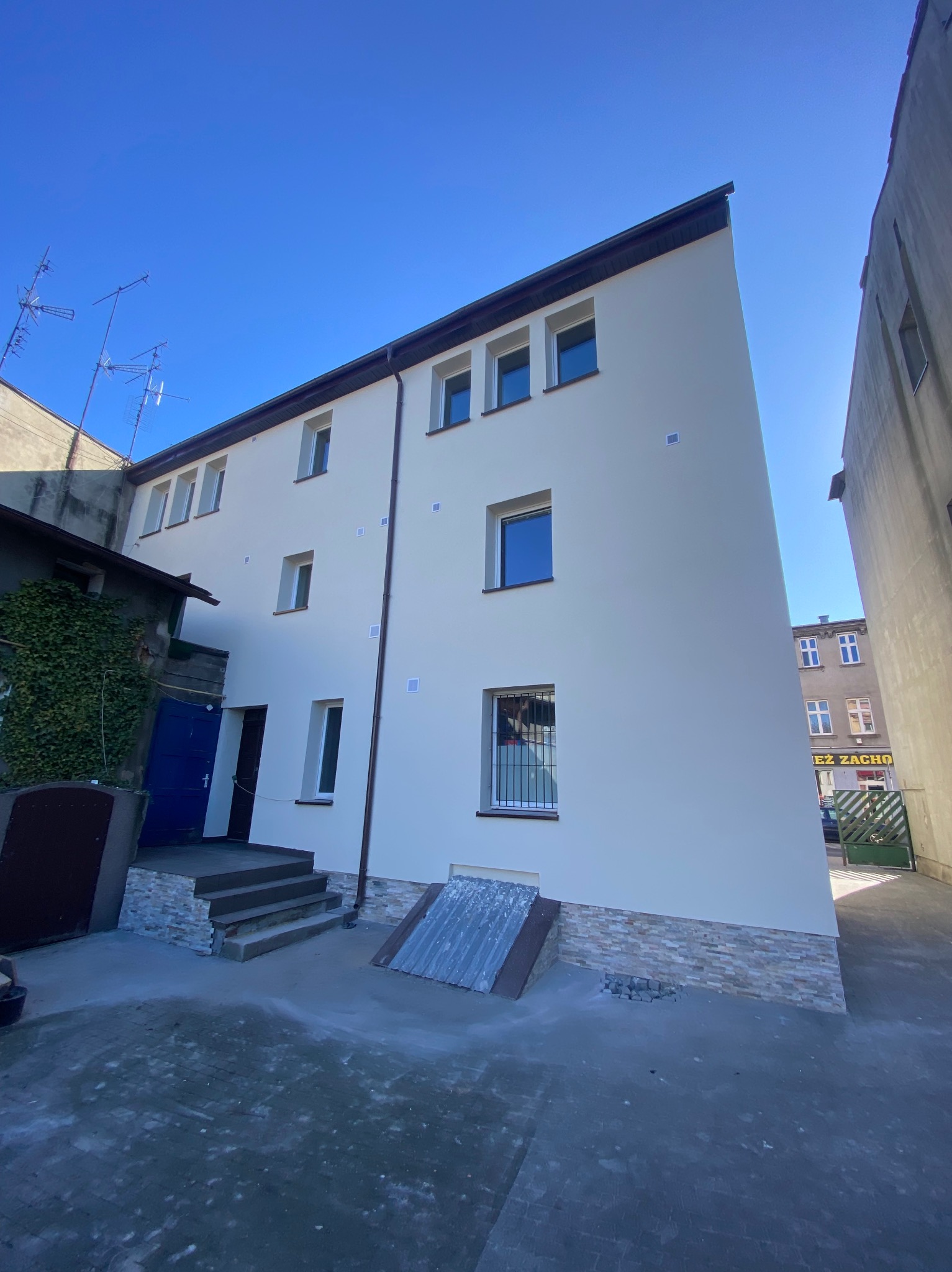 Odnowiona elewacja budynku z jasną fasadą, ciemnym dachem i nowymi oknami, widoczna z perspektywy podwórza w słoneczny dzień.