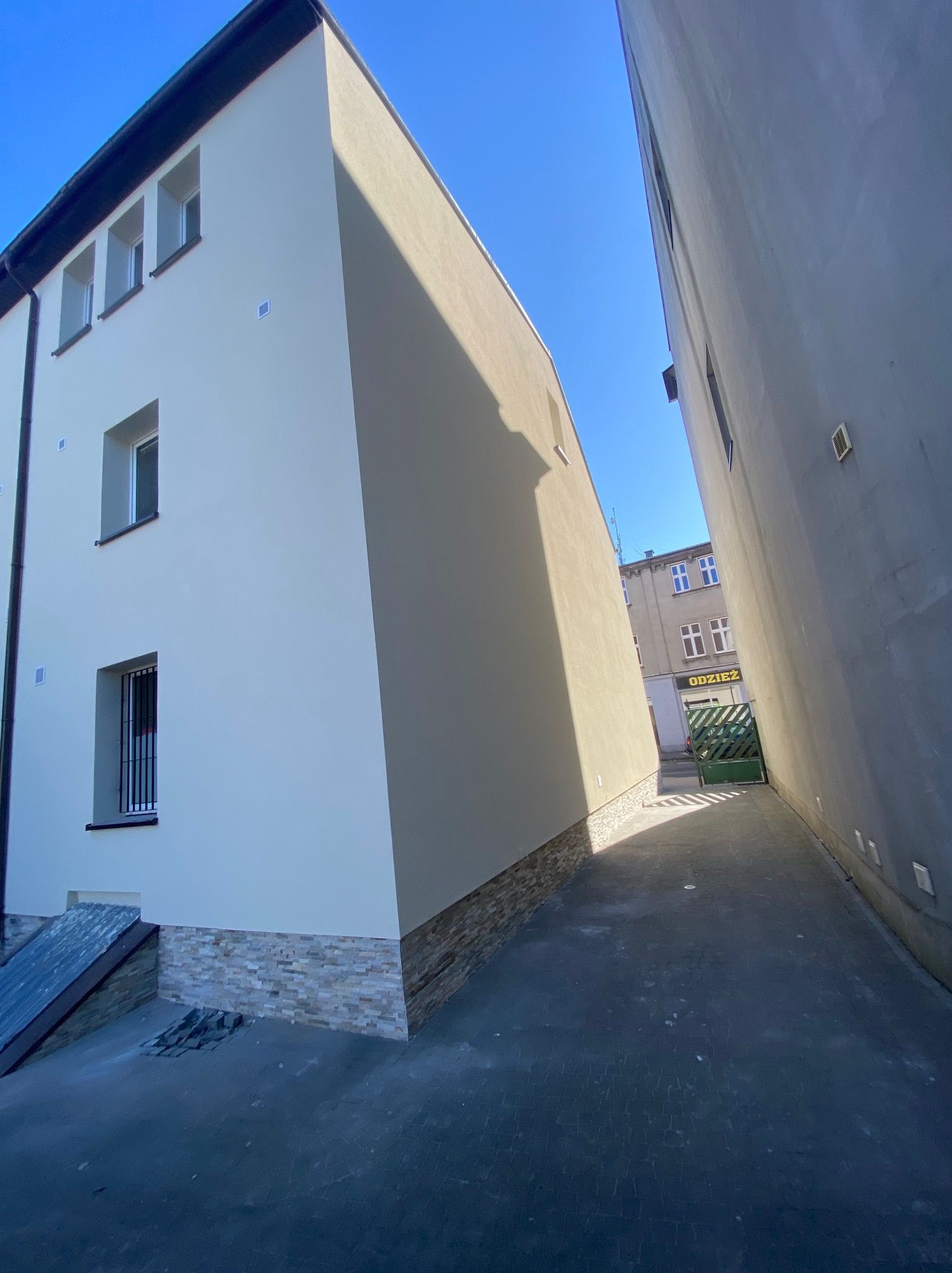 Elewacja po remoncie: budynek z jasną fasadą, wykończony kamieniem dekoracyjnym u podstawy, widziany z wąskiej uliczki w słoneczny dzień.