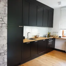 Nowoczesna kuchnia z czarnymi szafkami i drewnianym blatem, ściana z efektem starej cegły, okno z żaluzjami i widokiem na ulicę.