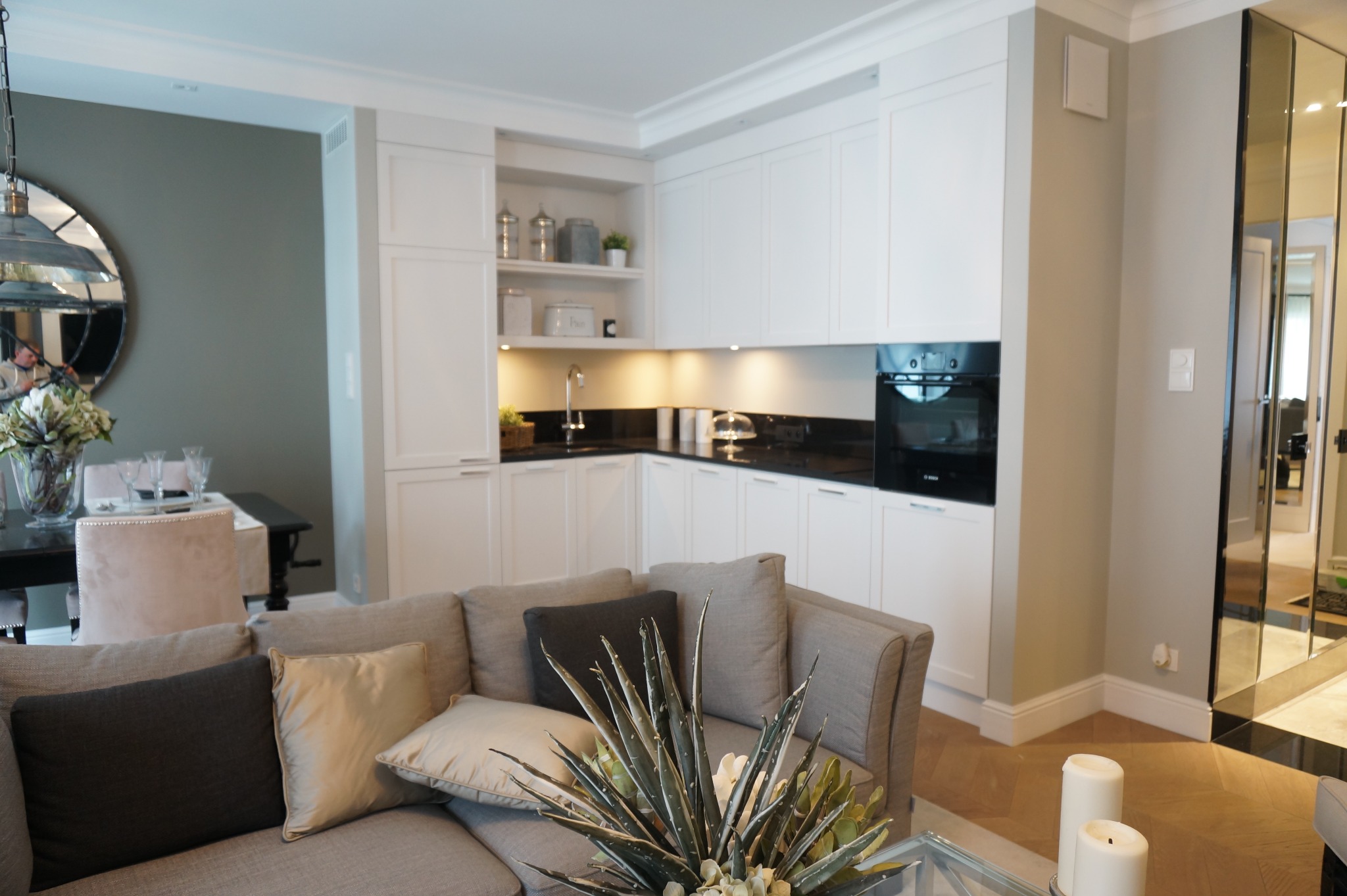 Jasne, nowoczesne wnętrze z białymi szafkami kuchennymi, czarnym blatem, zabudowaną piekarnikiem, szklanymi drzwiami i szarą kanapą z poduszkami oraz rośliną w centrum kadru.