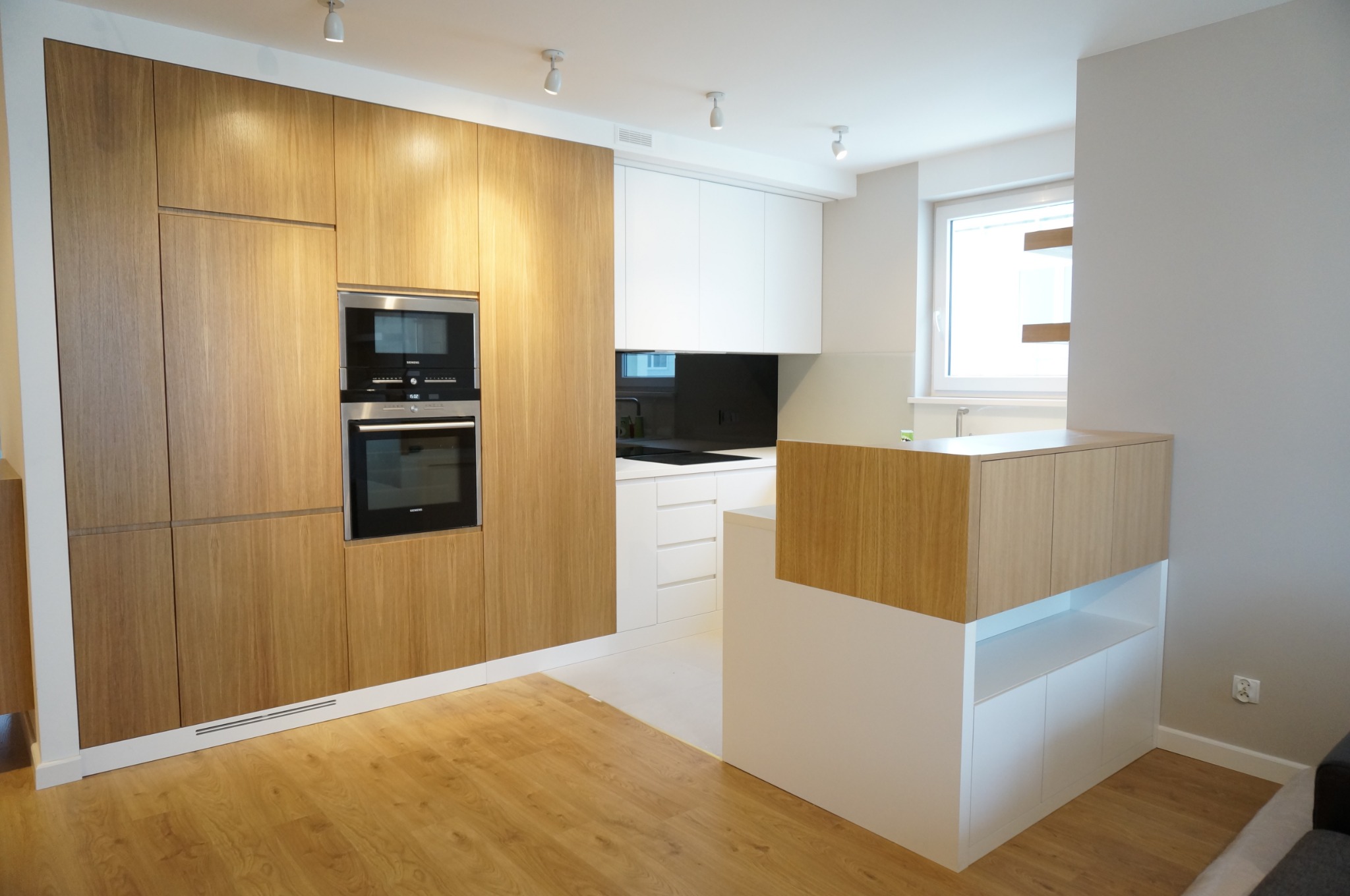 Nowoczesna kuchnia z drewnianymi frontami szafek i zabudowaną piekarnikiem oraz mikrofalówką, białe szafki wiszące i wyspa kuchenna z drewnianym blatem, jasna podłoga.