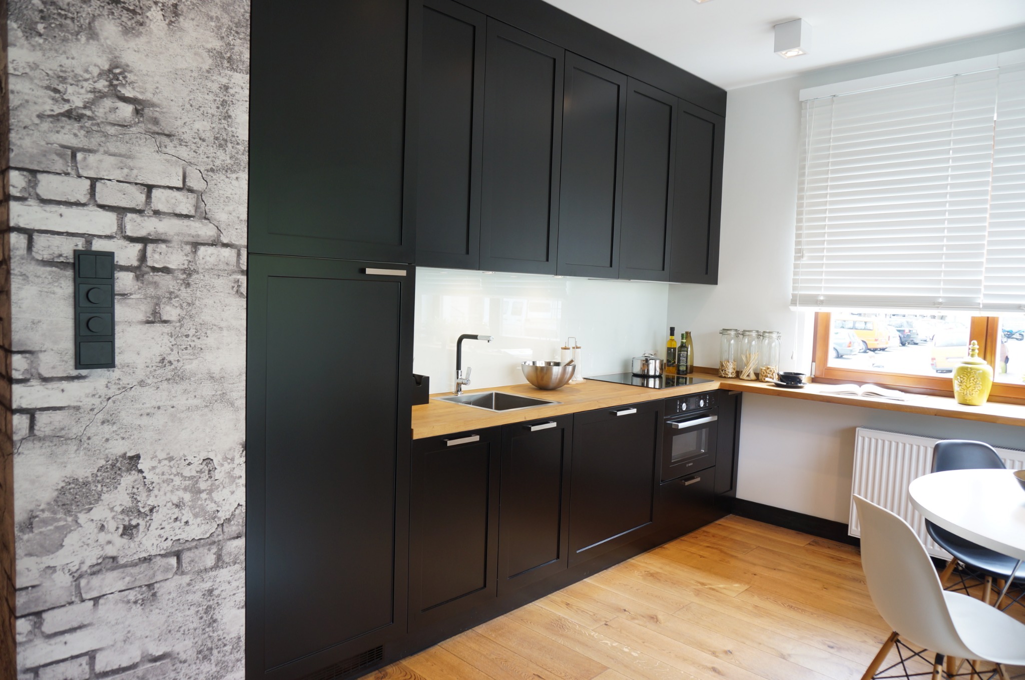 Nowoczesna kuchnia z czarnymi szafkami i drewnianym blatem, ściana z efektem starej cegły, okno z żaluzjami i widokiem na ulicę.