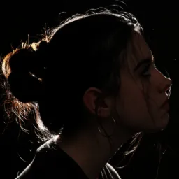 Profil młodej kobiety z włosami upiętymi w kok, oświetlonymi od tyłu, z kolczykami kołami na czarnym tle.