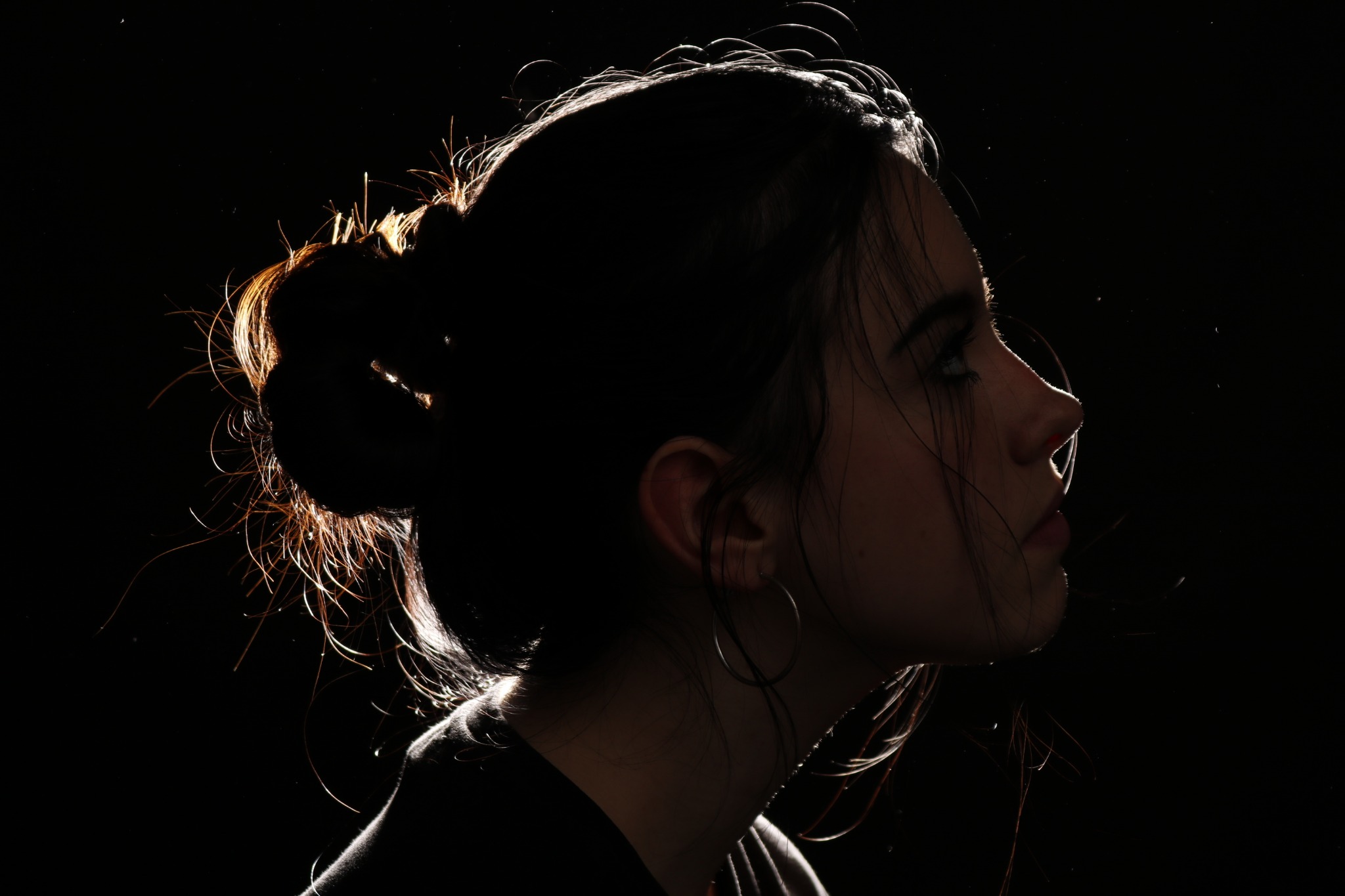 Profil młodej kobiety z włosami upiętymi w kok, oświetlonymi od tyłu, z kolczykami kołami na czarnym tle.