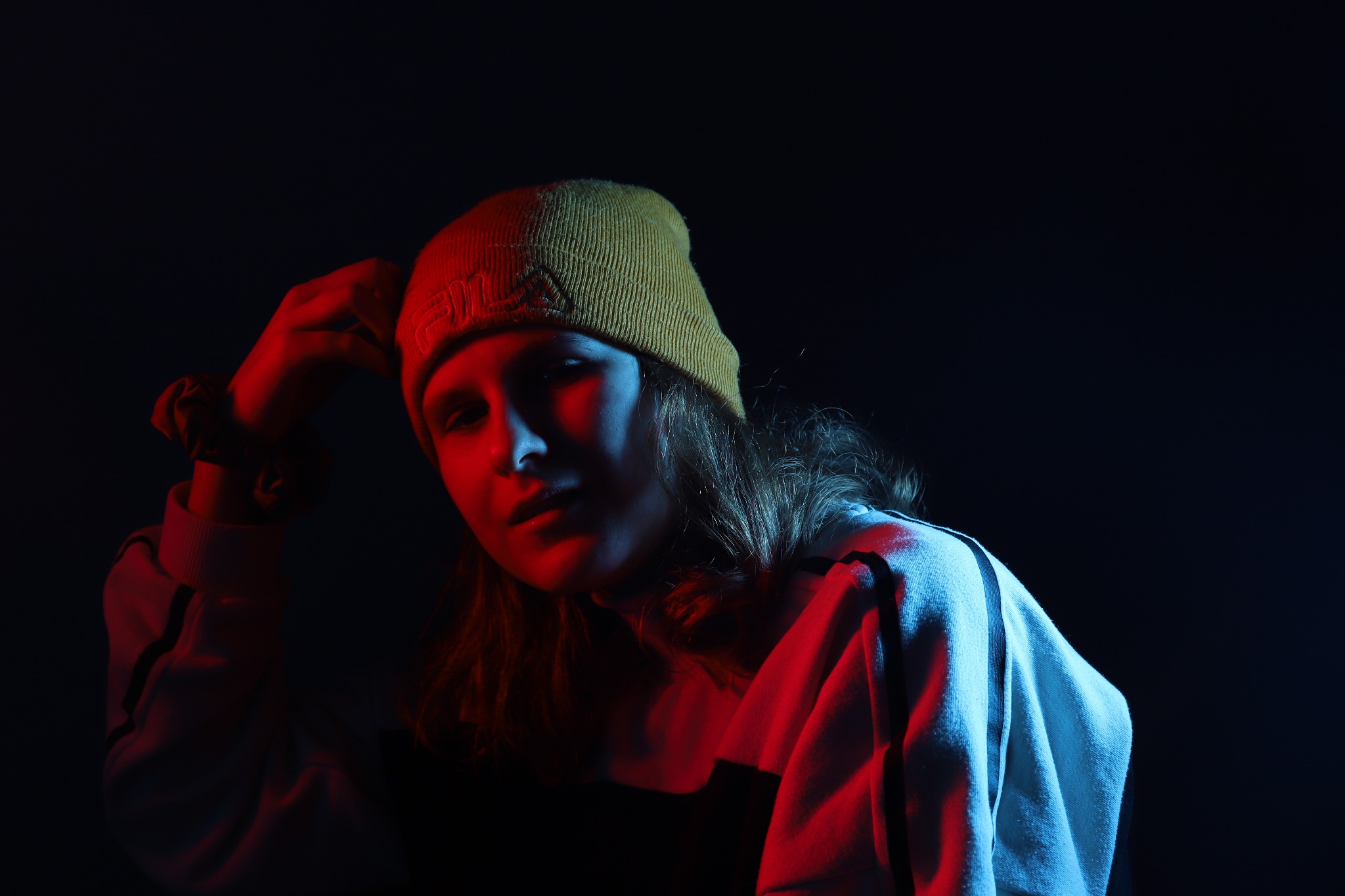 Portret kobiety w żółtej czapce Fila, oświetlonej czerwonym i niebieskim światłem, z dłonią uniesioną do głowy i gumką na nadgarstku.