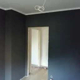 Ciemnoszara ściana w rogu pokoju z białym sufitem i listwą, widok na jasne pomieszczenie przez framugę drzwi, widoczne kable wystające z sufitu.