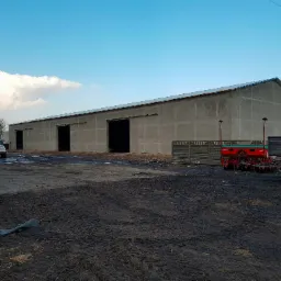 Szeroki, parterowy budynek gospodarczy o szarych ścianach z trzema otwartymi wrotami, na terenie budowy z błotnistym podłożem, z zaparkowanymi pojazdami i sprzętem rolniczym.