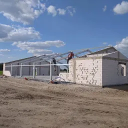 Wznoszenie ścian budynku z białych bloczków, widoczne stalowe elementy konstrukcji dachu, dwóch robotników przy pracy, jeden na drabinie, drugi siedzi na ścianie, w tle pole i błękitne niebo...