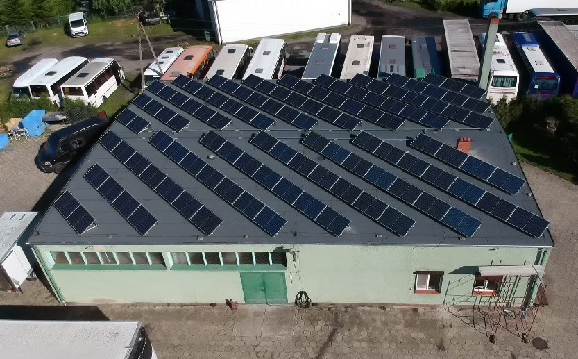 Widok z góry na halę z panelami słonecznymi na dachu, zaparkowane autobusy w tle, ceglany komin.