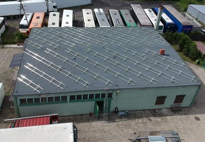 Hala przemysłowa z zamontowaną konstrukcją pod panele fotowoltaiczne, widok z góry, na dachu ceglany komin, w tle zaparkowane ciężarówki i autobusy.