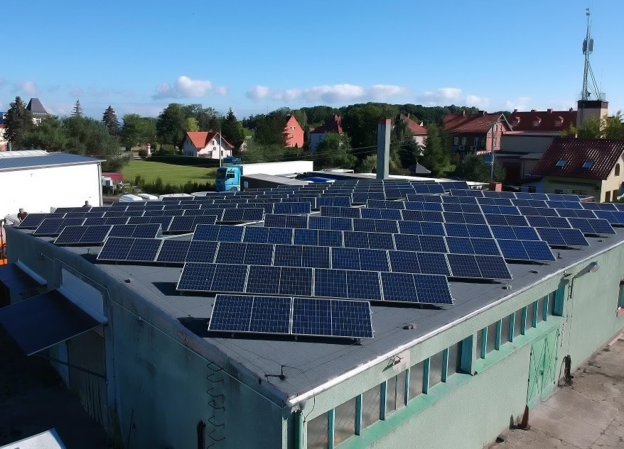 Panel słoneczny zamontowany na dachu hali produkcyjnej, widok z góry, w tle zabudowania mieszkalne i niebieska ciężarówka.