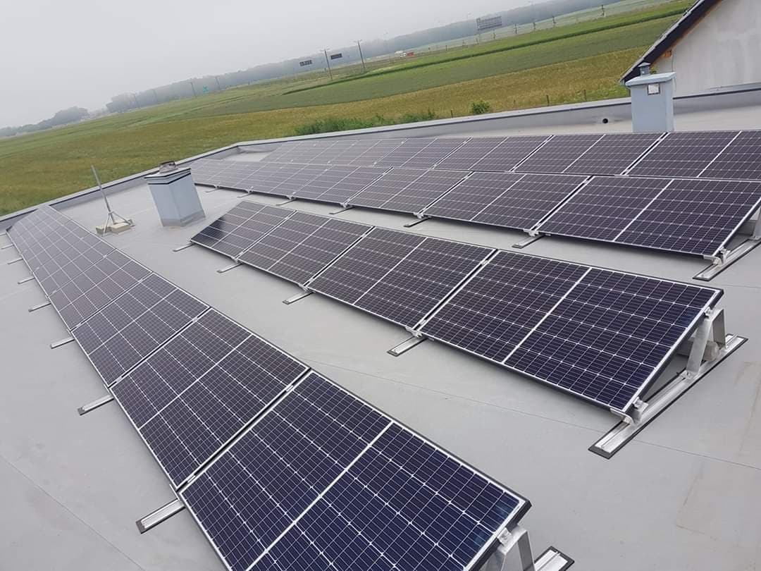 Montaż paneli słonecznych na dachu hali przemysłowej, widok z góry, w tle pola uprawne i zamglone niebo.