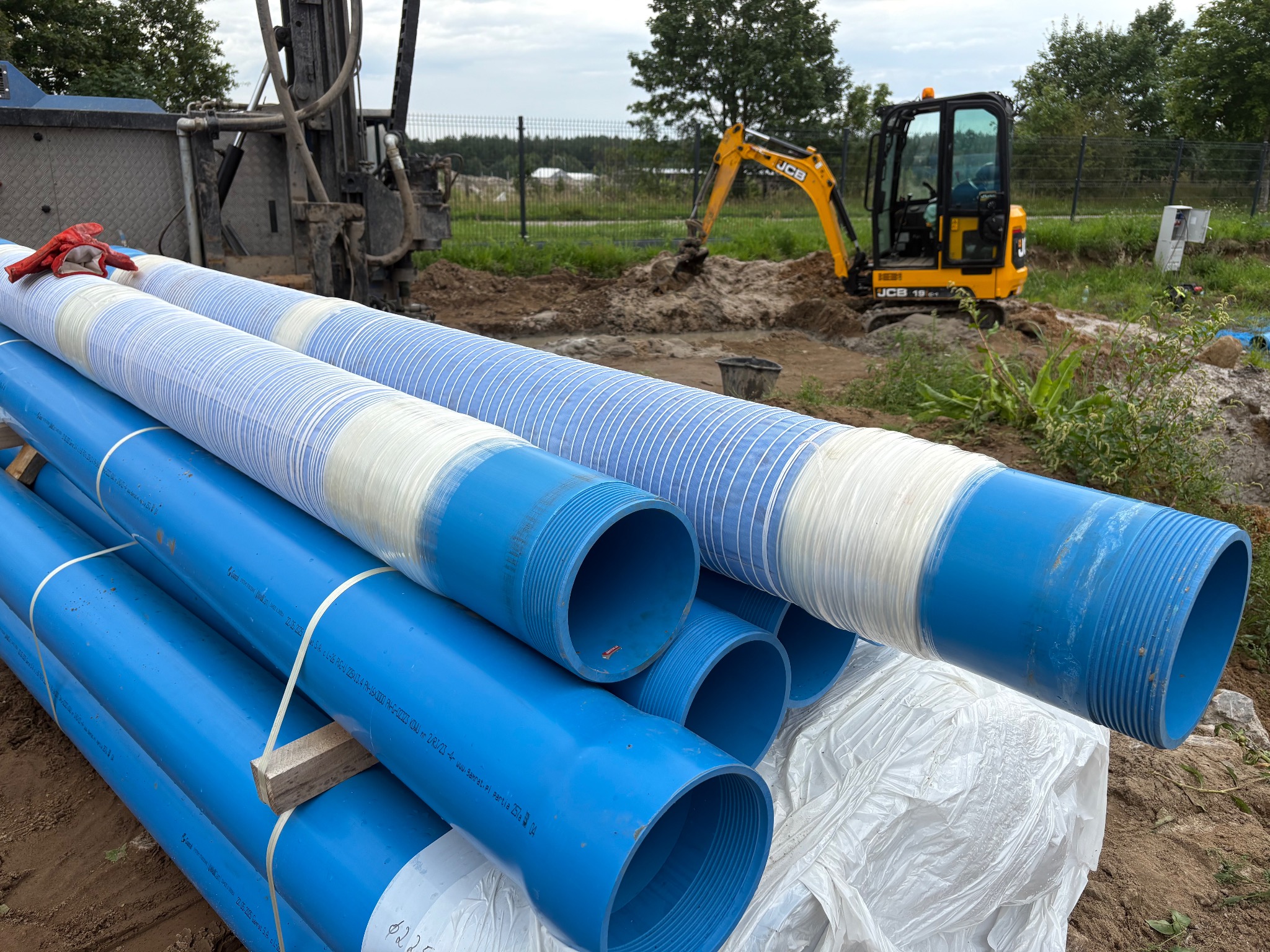 Niebieskie rury studzienne z gwintem na placu budowy, w tle koparka JCB i wiertnica. Widoczne rękawice robocze na rurach.