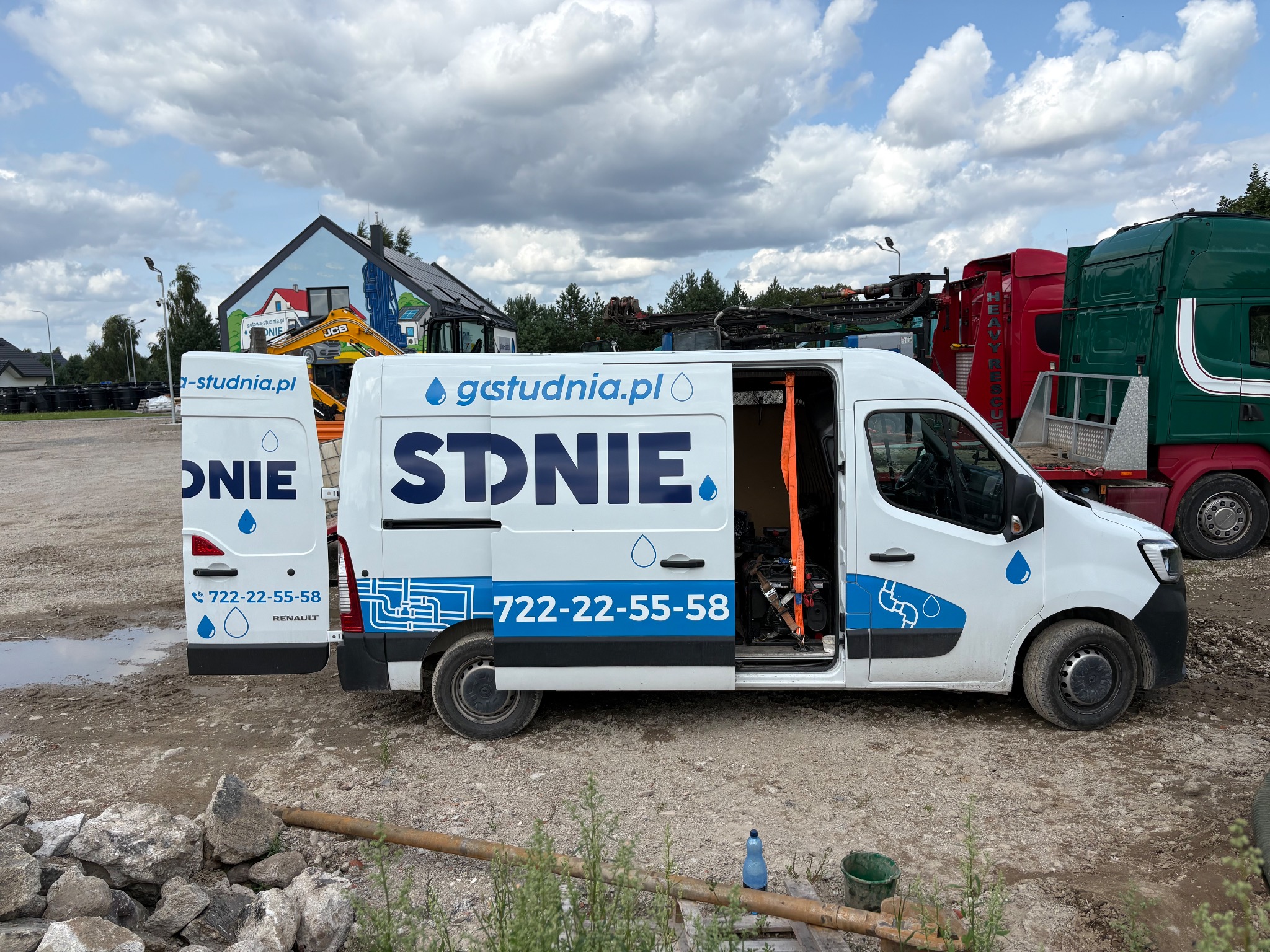 Biały van z logo firmy studniarskiej na placu budowy, otwarte drzwi boczne i tylne, widoczne narzędzia i sprzęt. W tle budynek z muralem i ciężarówki.