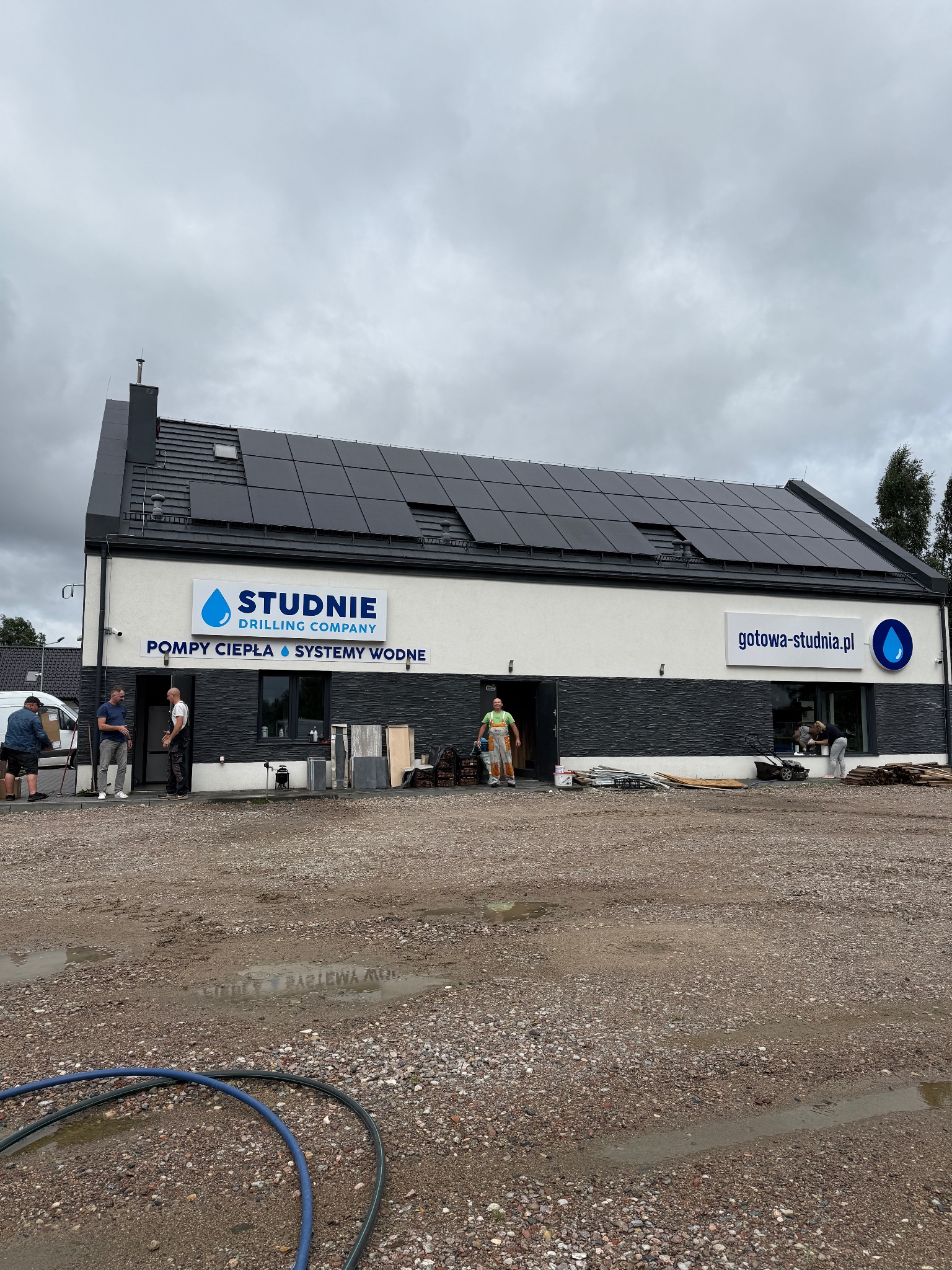 Budynek firmy Studnie Drilling Company z panelami słonecznymi na dachu, oferującej pompy ciepła i systemy wodne. Na zewnątrz pracują osoby, widoczne materiały budowlane.