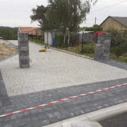 Świeżo ułożona, jasnoszara kostka brukowa z ciemnoszarą obwódką, ograniczona słupkami z szarego kamienia i taśmą ostrzegawczą, z ubijarką w tle.