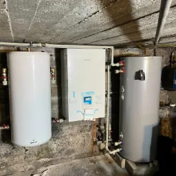 Zestaw urządzeń grzewczych: biały zbiornik Galmet, pompa ciepła Panasonic i szary zbiornik, zainstalowane w pomieszczeniu z surowym betonowym stropem.